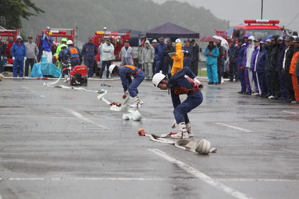 雨の中、消防団員が、地面に置いてあるホースを拾おうとしている写真